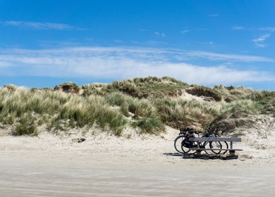 Zwei Fahrräder, die vor einer großen Sanddüne mit Gräsern und Schilf unter blauem Himmel abgestellt sind